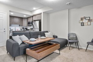 Una sala de estar moderna con un sofá gris, una mesa de centro de madera y una cocina al fondo.