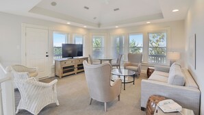 Un salon spacieux doté d’un ventilateur de plafond, de grandes fenêtres, d’un téléviseur à écran plat, de meubles en osier et d’une table basse en verre.