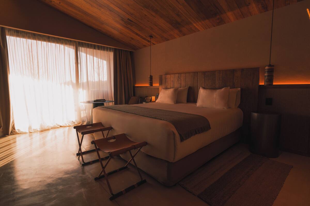 A modern bedroom with a large bed, wooden ceiling, and a view of a cityscape through large windows.