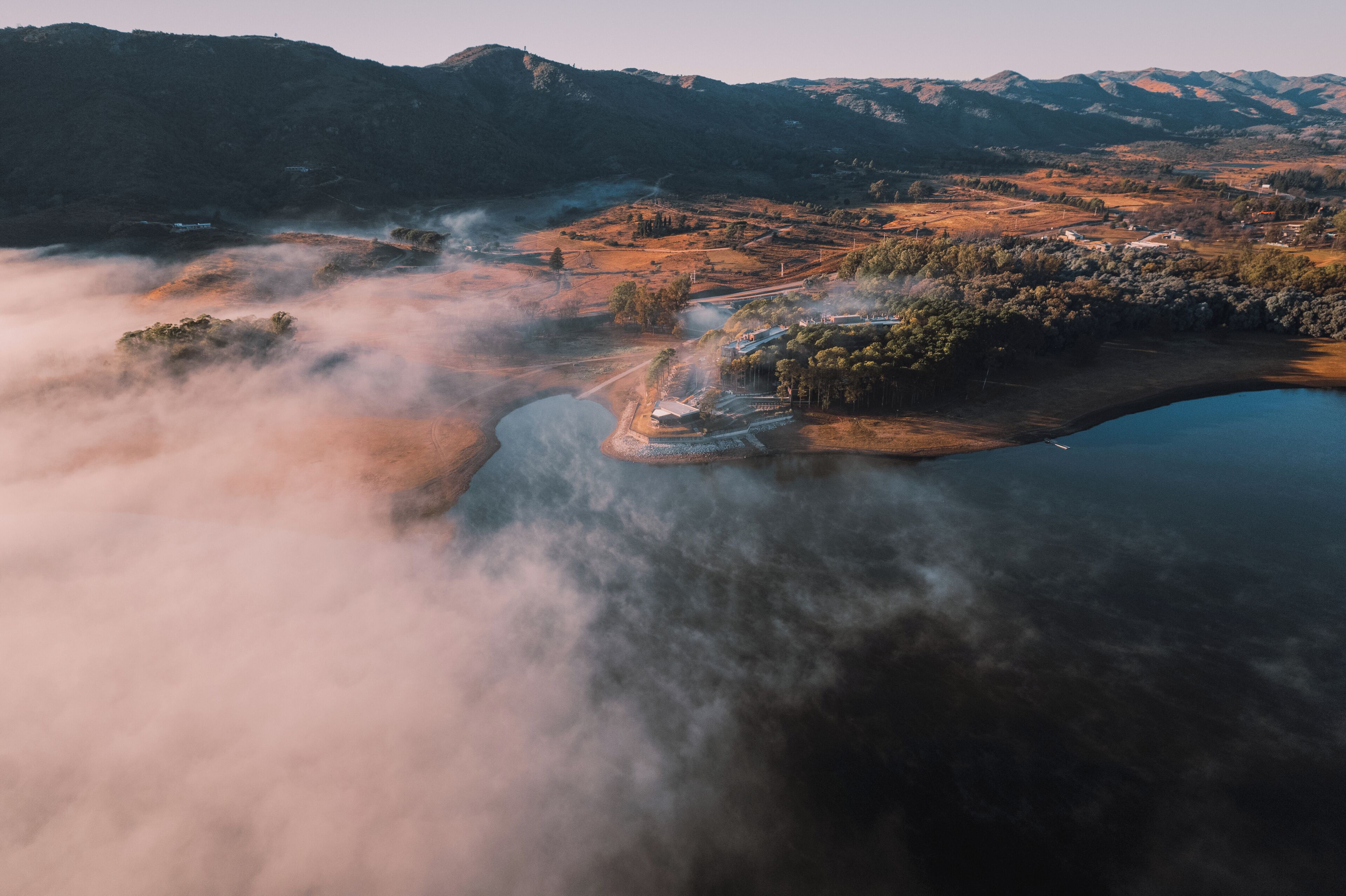 Vista aérea de un valle con un lago, rodeado de montañas y un ambiente brumoso.