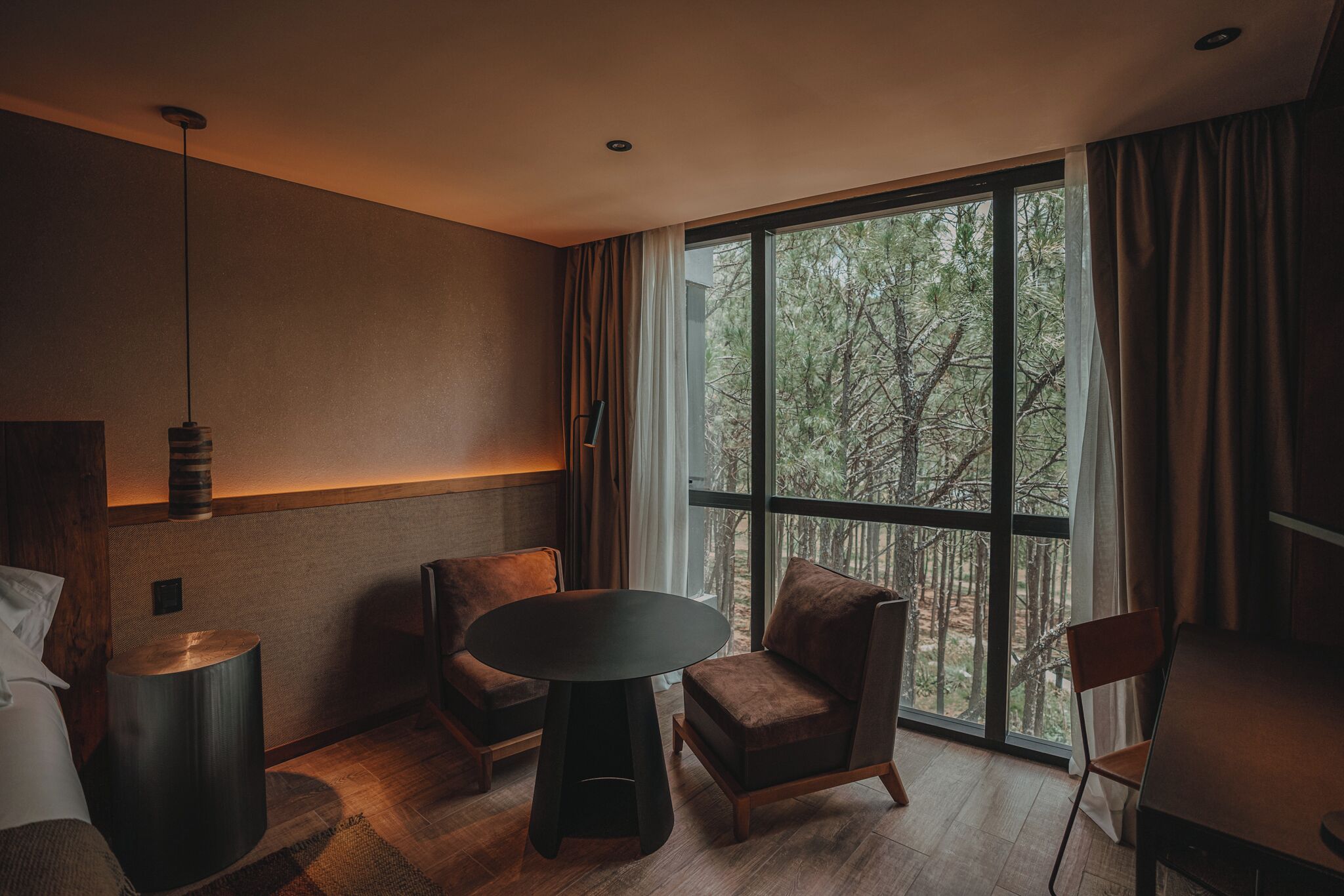 A modern room with a large window, a round table, two chairs, a bed, and a lamp.