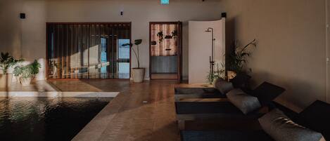 A modern indoor pool area with a wooden floor, seating area with cushions, and potted plants.