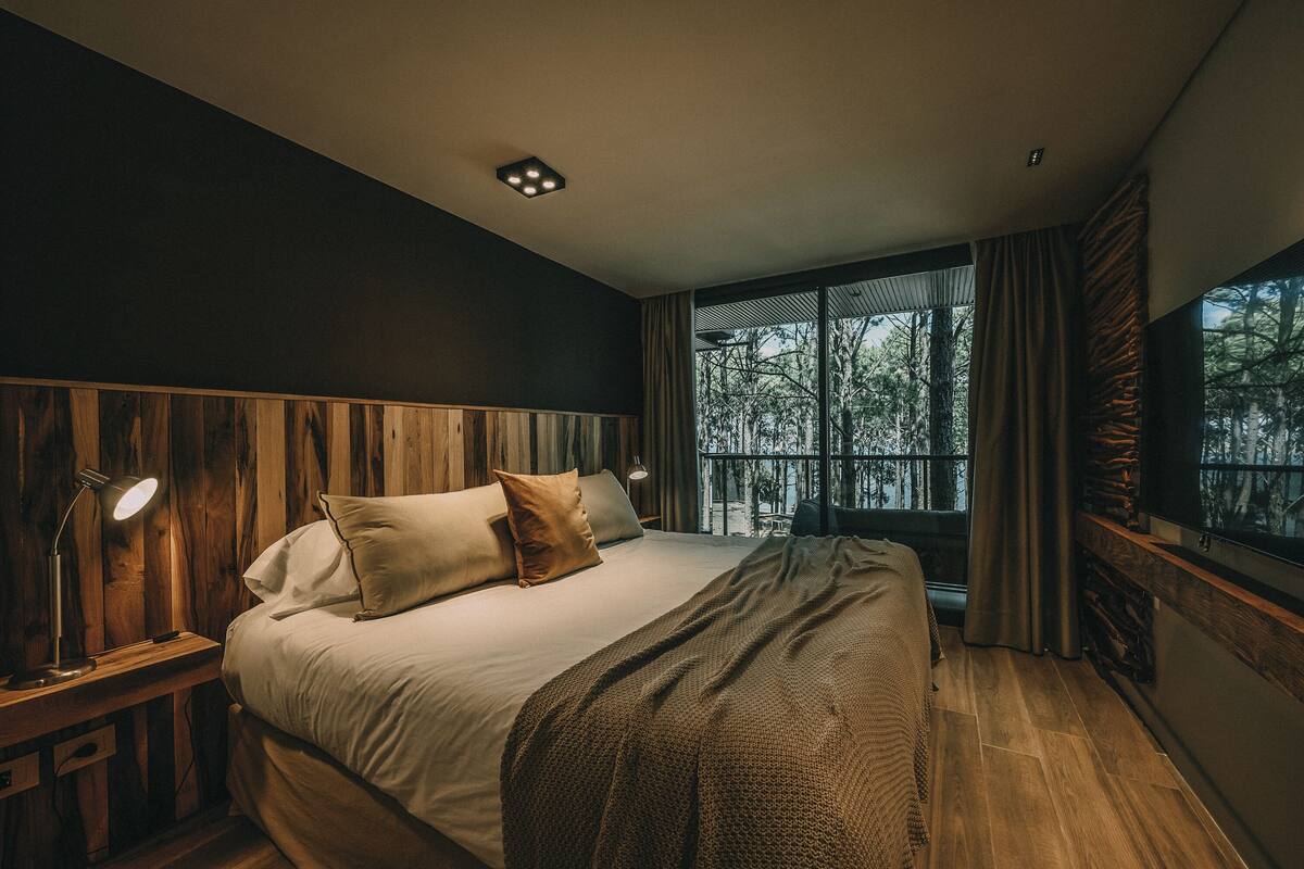 A bedroom with a large bed, wooden headboard, a nightstand with a lamp, and a window with curtains.