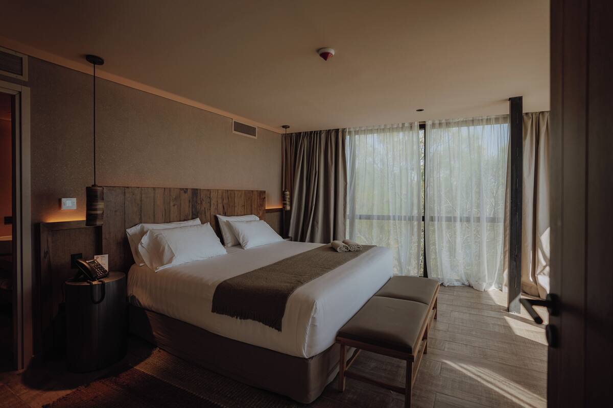 A hotel room with a large bed, a bedside table with a phone, a bench, and a view of the outdoors through sheer curtains.