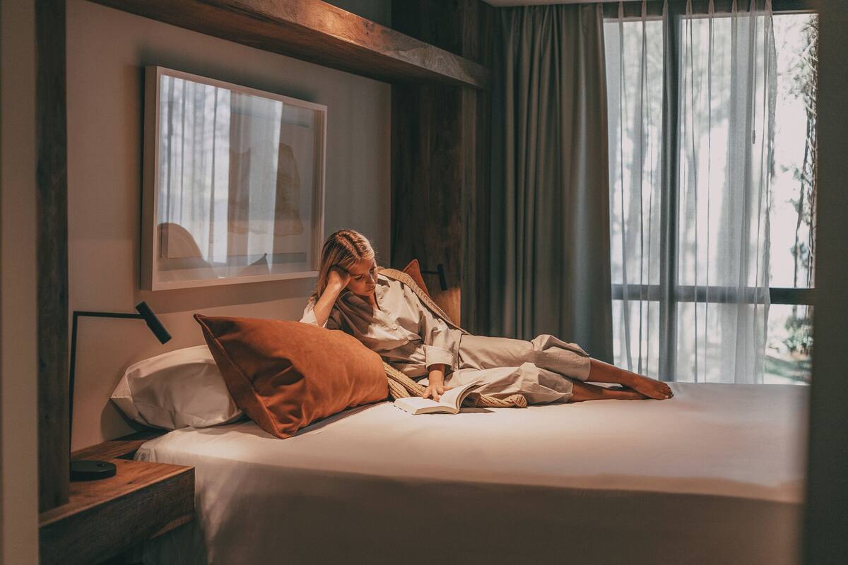 A bedroom with a bed, pillows, a bedside table, a lamp, and a window with curtains.