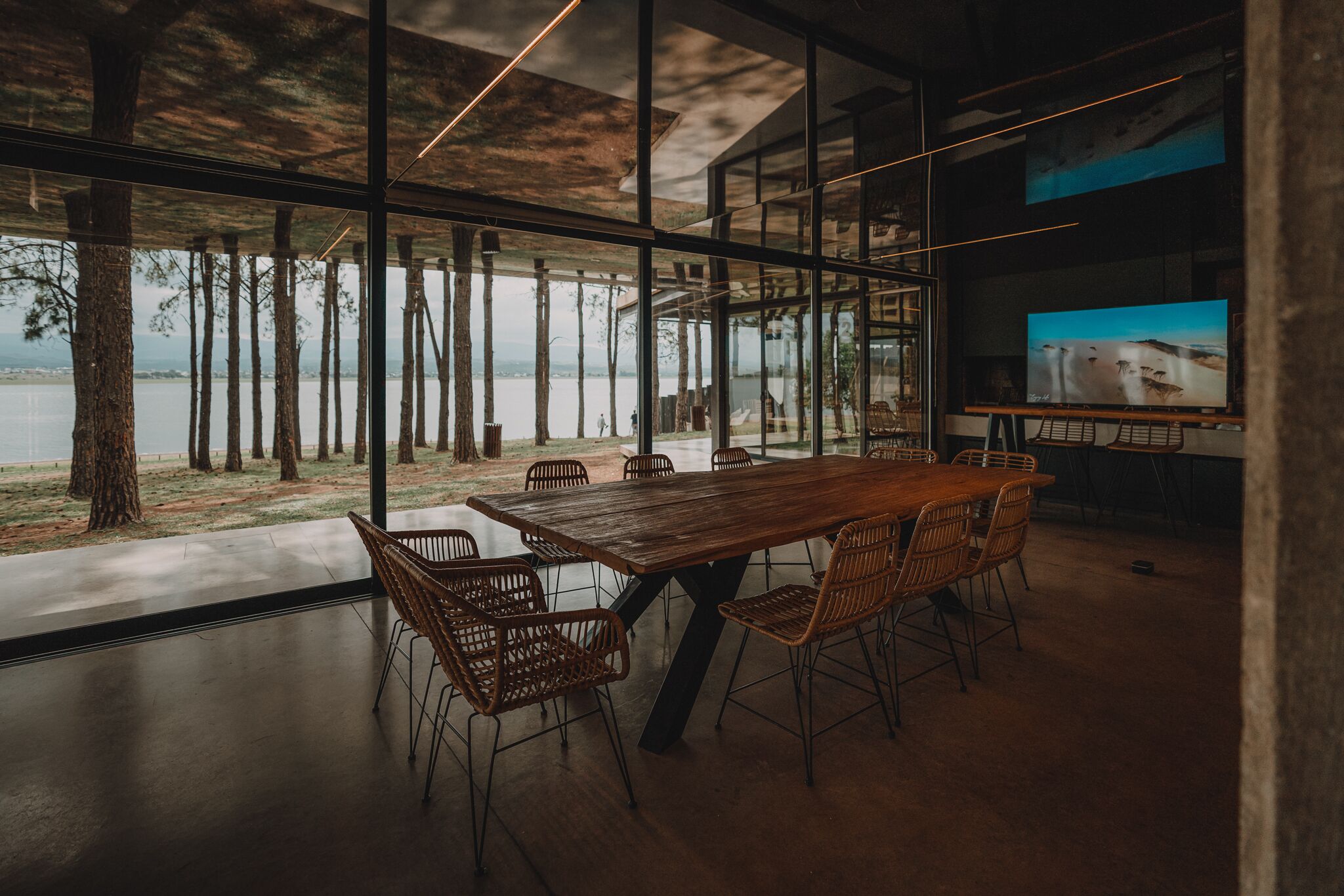 Un espace repas moderne avec une grande table en bois et des chaises en osier, de grandes fenêtres offrant une vue sur les arbres et un plan d’eau, et un téléviseur à écran plat fixé au mur.