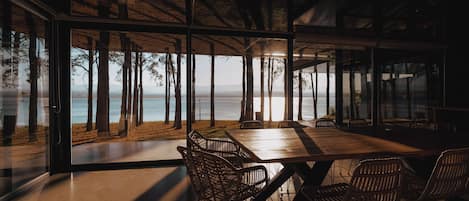 A modern dining area with a large table and wicker chairs, overlooking a beach and the sea.