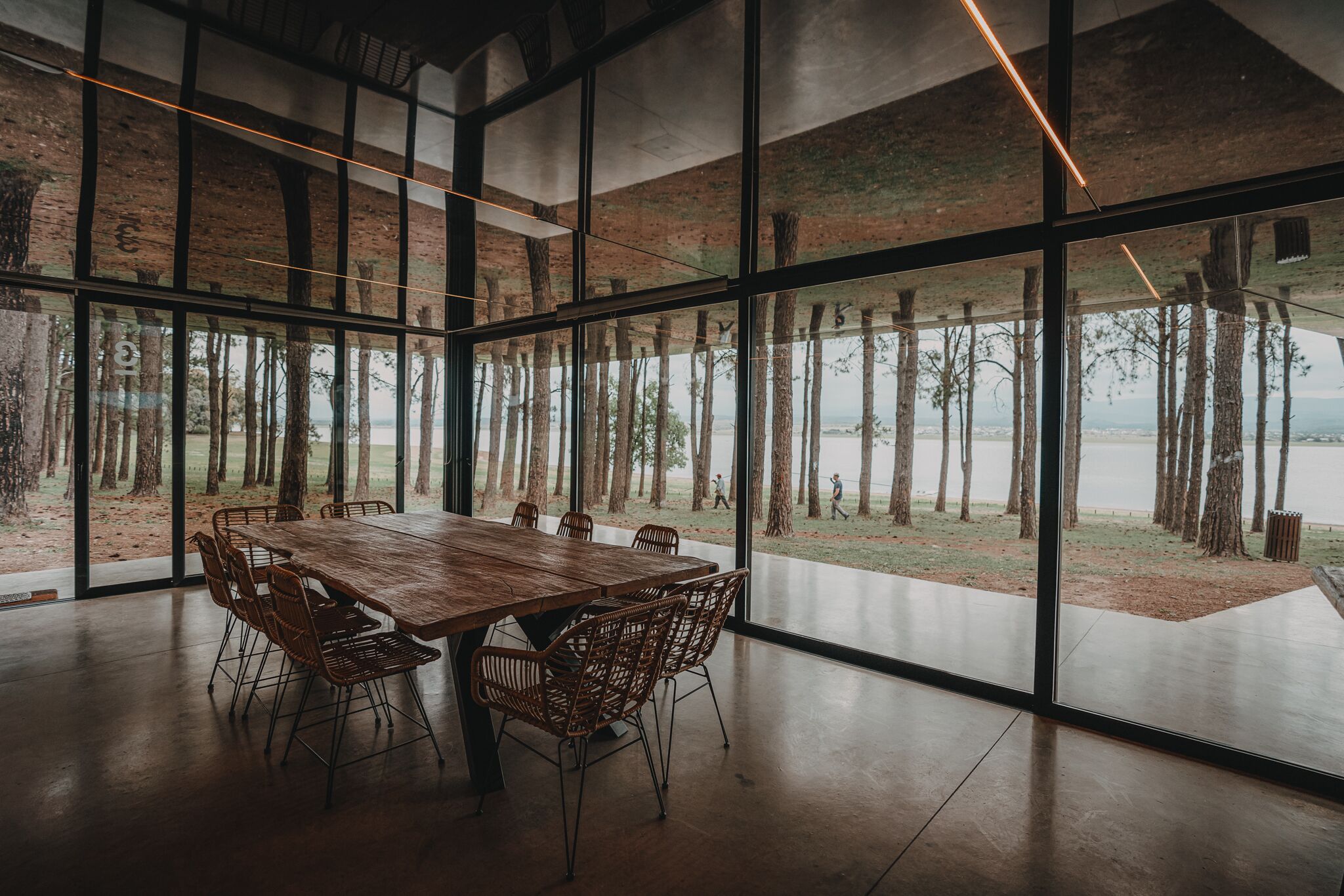 Una habitación amplia con una gran mesa de madera y sillas de mimbre, ventanas grandes que ofrecen vistas a árboles y un paisaje lejano.