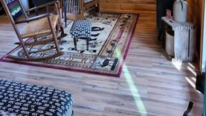 Ein gemütliches Blockhaus-Interieur mit Holzwänden, Deckenventilator, Kühlschrank, Mikrowelle, einem gemusterten Teppich, einem Schaukelstuhl, einem Tisch mit Lampe und einem Fenster mit Blick.
