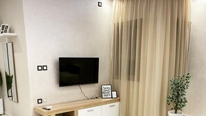 Un salon moderne avec un téléviseur à écran plat fixé au mur, un meuble de rangement en bois, une table à manger avec des chaises et une grande fenêtre ornée de rideaux beiges.