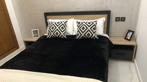 Un lit recouvert d’une couverture et de coussins à motifs noir et blanc, une table de chevet avec un cadre photo, et une petite table de nuit également ornée d’un cadre photo.