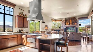 A spacious kitchen with a large island, stone fireplace, and a dining area with a view of the outdoors. - Gorgeous Desert Home W/ Backyard Oasis! (Cave Creek)