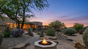A landscaped outdoor area with a fire pit, cacti, and a building with large windows. - Gorgeous Desert Home W/ Backyard Oasis! (Cave Creek)