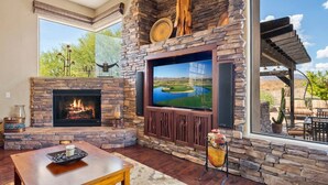 A living room with a stone fireplace, a large flat-screen TV, and a view of a golf course through a large window. - Gorgeous Desert Home W/ Backyard Oasis! (Cave Creek)
