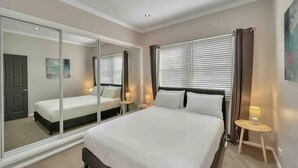 A bedroom with a large bed, a mirror, a window with blinds, and a small bedside table with a lamp.