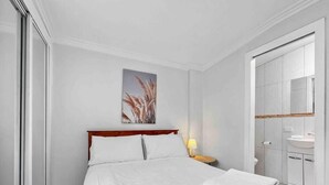 A hotel room with a bed, a nightstand with a lamp, a bathroom with a sink and mirror, and a painting on the wall. - Apt 3 Brightside Apartments - City Center (Armidale)