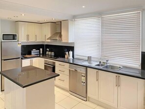 A modern kitchen with white cabinets, stainless steel appliances, a black countertop, and a window with blinds. - Apt 1 Brightside Apartments - City Centre (Armidale)