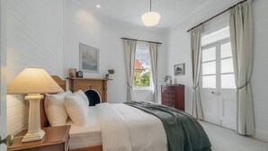 Ein Schlafzimmer mit Bett, Nachttisch, Lampe, Kommode und einem Fenster mit Vorhängen.
