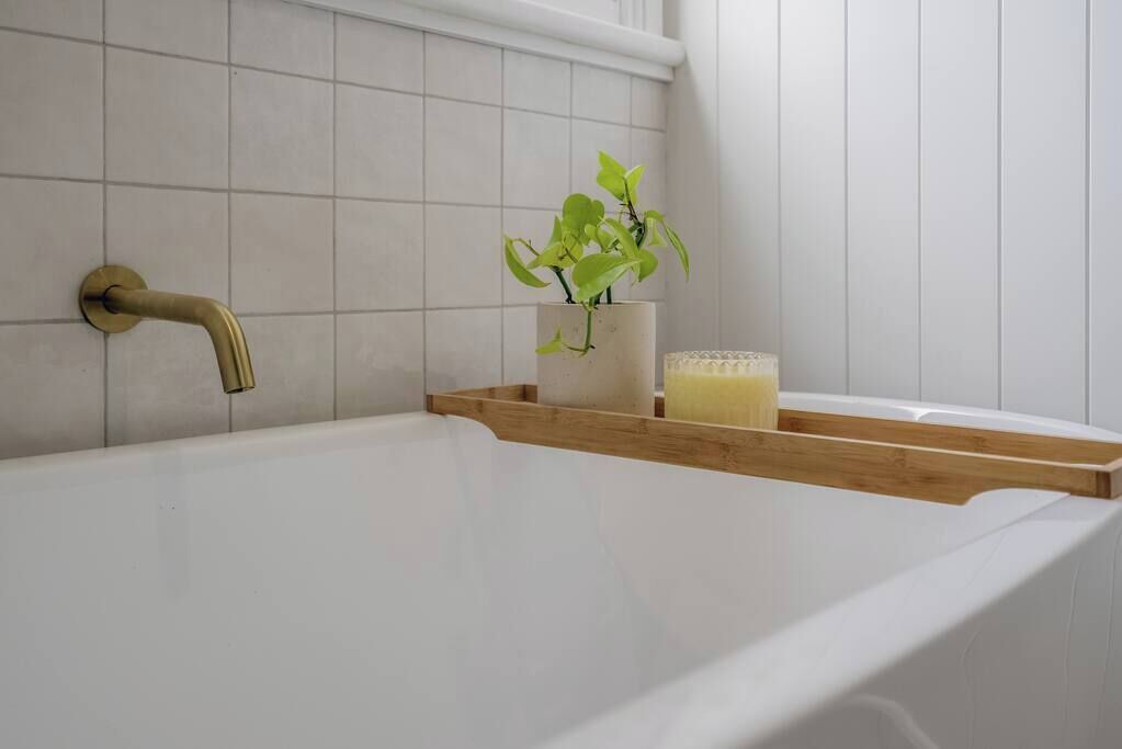 Ein Badezimmer mit einer weißen Badewanne, einem goldenen Wasserhahn, einem Holzregal, einer Topfpflanze und einer Kerze.