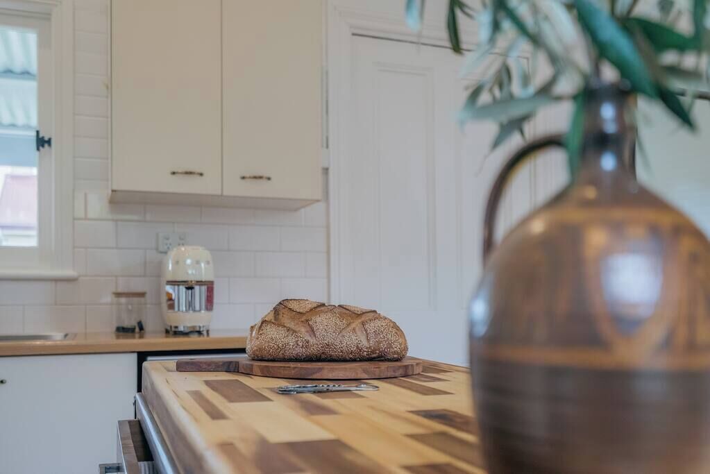 Een keuken met witte kastjes, een brood op een houten snijplank, en een vaas met een plant.