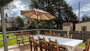 Eine Holzterrasse mit einem weißen Tisch und Stühlen, einem Sonnenschirm und Blick auf einen Garten mit Bäumen und einem Haus.