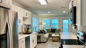 A modern kitchen with a refrigerator, stove, and microwave. There is a dining area with a table and chairs, and a sofa. The room has large windows and a ceiling light. - Sublime Sunset☀️ Ocean Front Getaway (Port St. Joe)