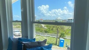 A room with a large window overlooking a beach and a road. There are several blue and striped pillows on a bench. - Sublime Sunset☀️ Ocean Front Getaway (Port St. Joe)