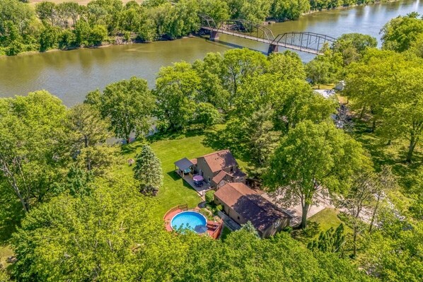 An aerial view of a residential property with a swimming pool, a house, and a bridge over a river.