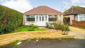 Une maison de plain-pied avec un toit recouvert de tuiles rouges, un porche à l’avant et un jardin délimité par une clôture en bois.