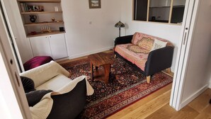 Ein gemütliches Wohnzimmer mit einer Couch, einem Couchtisch und einem gemusterten Teppich. Ein Bücherregal mit Dekorationselementen und ein gerahmtes Bild sind an der Wand zu sehen.