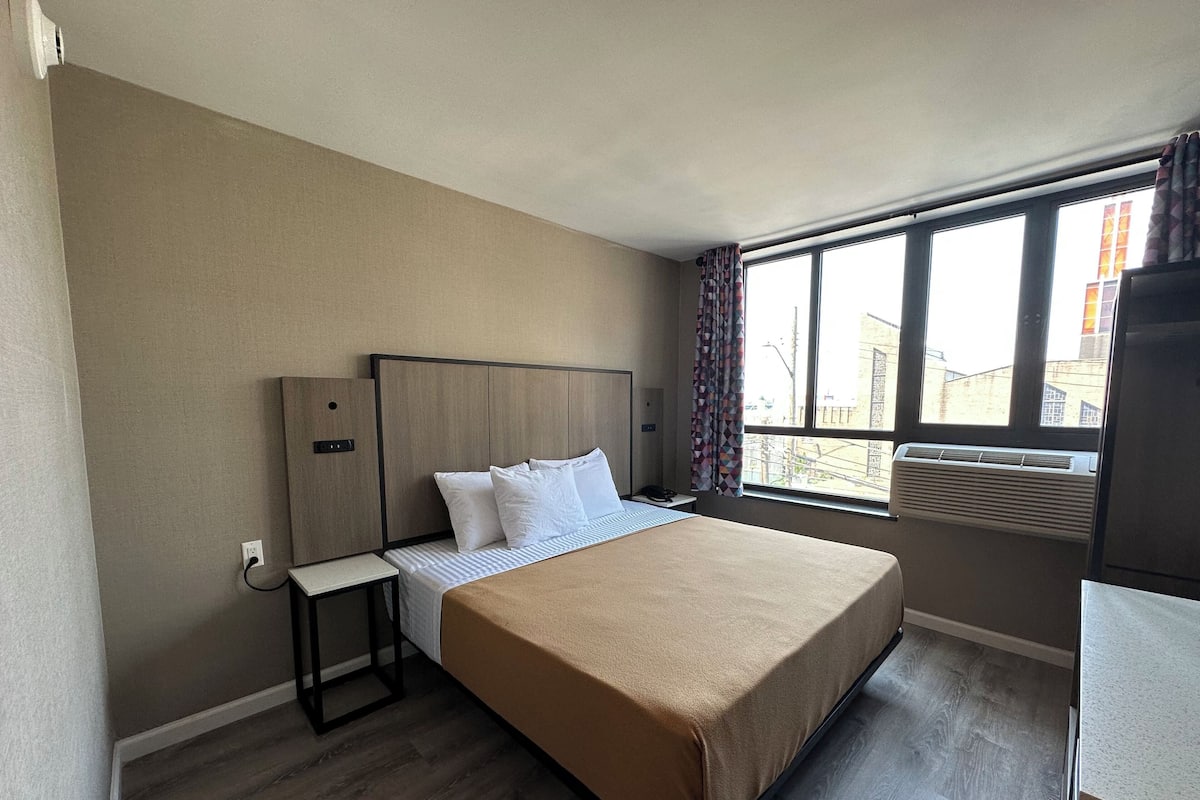 A hotel room with a bed, a bedside table, a window with curtains, and an air conditioning unit.