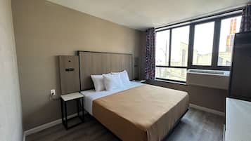 A hotel room with a bed, a bedside table, a window with curtains, and an air conditioning unit.