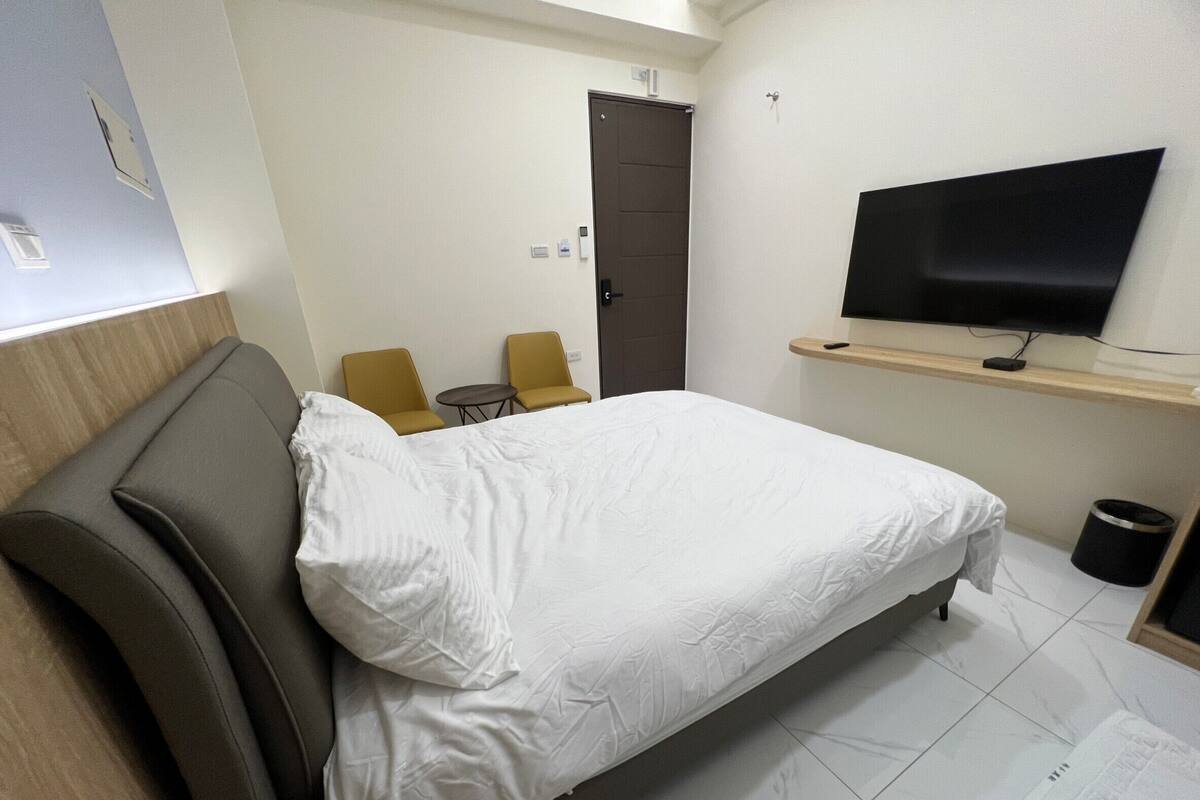 A hotel room with a bed, two chairs, a TV, and a door.