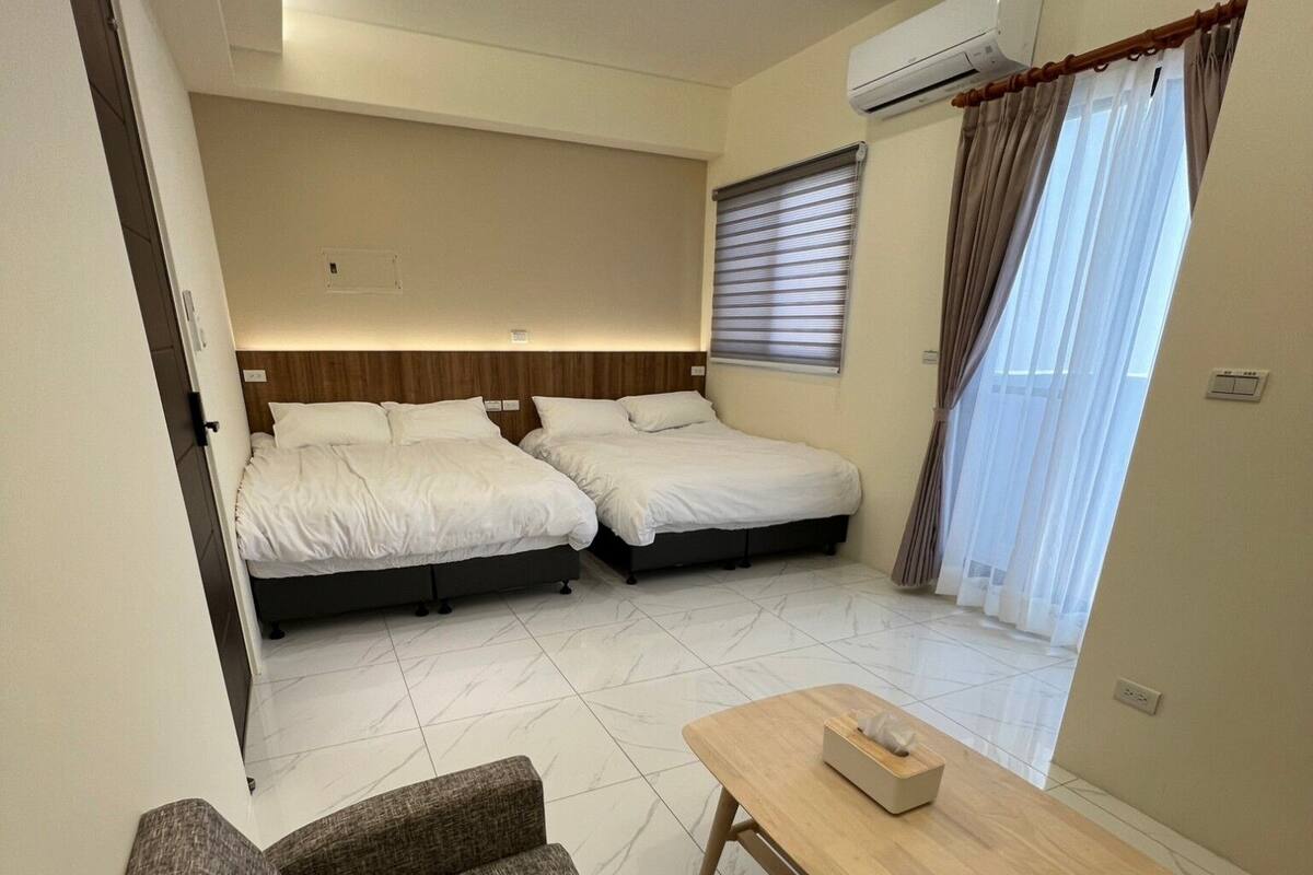 A hotel room with two beds, a sofa, a wooden table, and an air conditioning unit.