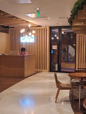 Un espace d’accueil avec un comptoir de réception en bois, un panneau néon, un lustre et une porte en verre donnant accès à une aire de jeux.