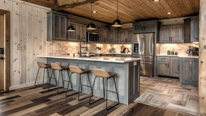 Une cuisine avec des meubles en bois, un îlot central doté de tabourets de bar, un réfrigérateur en acier inoxydable et des luminaires suspendus.