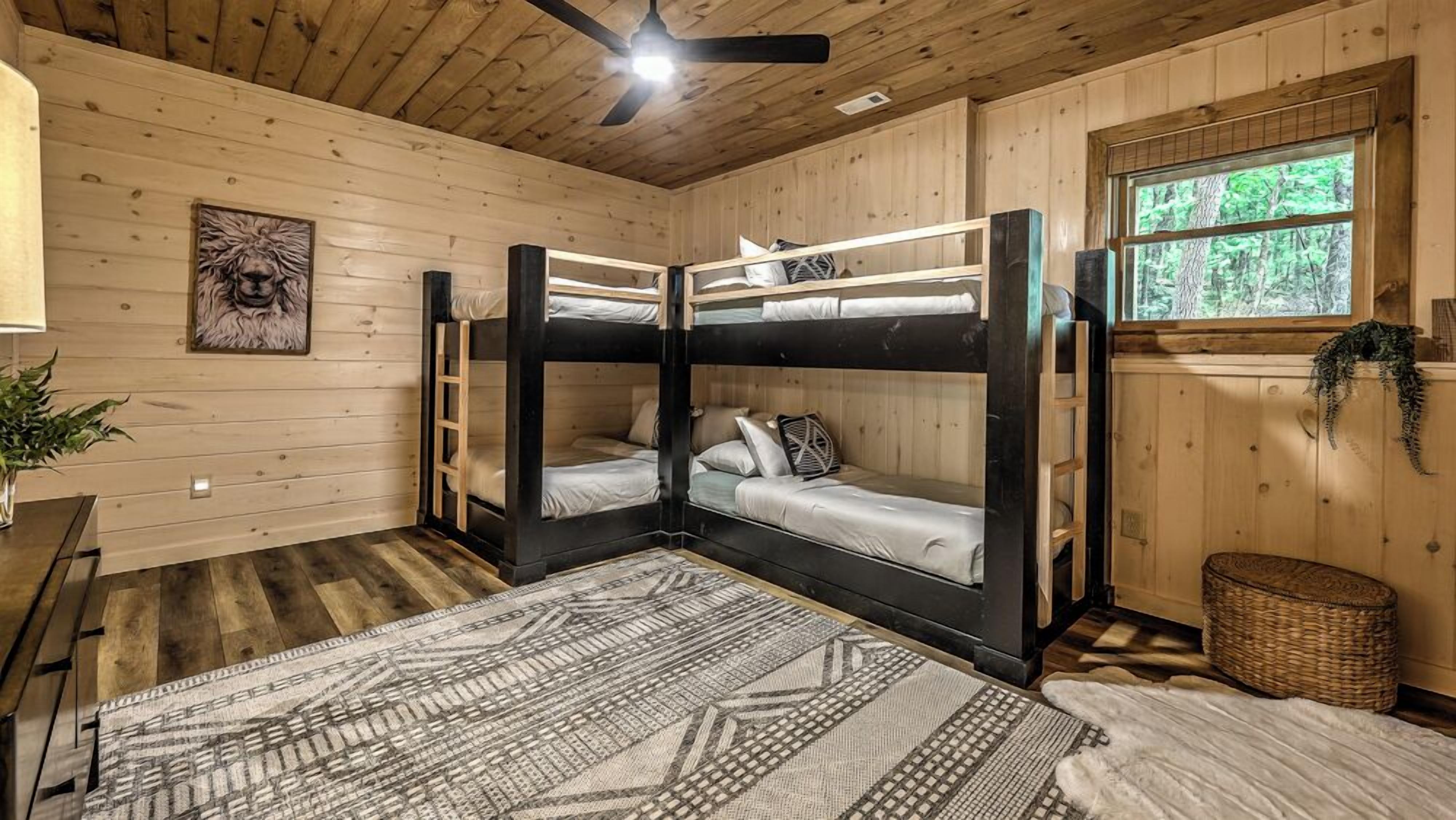 A bunk bed room with a ceiling fan, a window with a view of trees, a wooden floor, and a patterned rug.