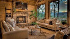 L’intérieur chaleureux d’un chalet avec une cheminée en pierre, un canapé et une grande fenêtre donnant sur la forêt.