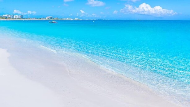 Een strand met helder turquoise water, wit zand en een zeilboot in de verte.