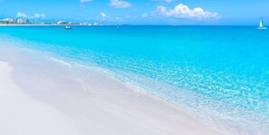 A beach with clear turquoise water, white sand, and a sailboat in the distance.