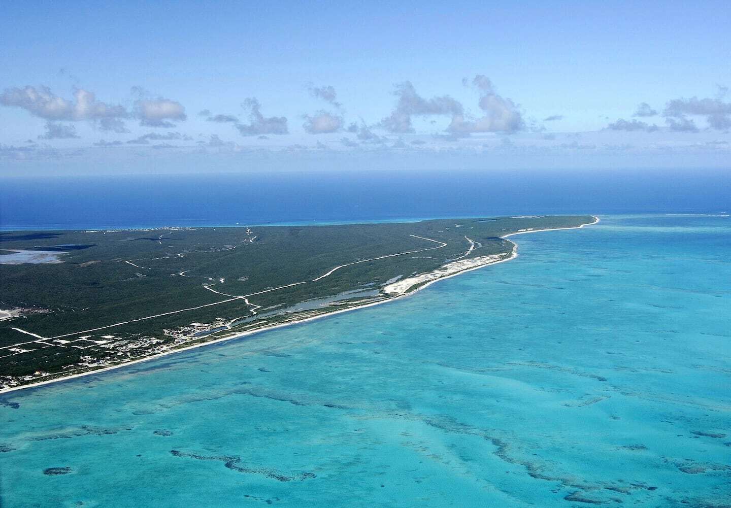 Luchtbeeld van een kustgebied met helder turquoise water, een smalle strook land met gebouwen en een dicht bebost gebied.