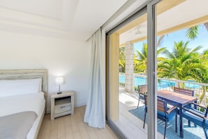 Habitación de hotel con cama, mesita de noche, lámpara y puerta corrediza de vidrio que da a un balcón con piscina y palmeras.