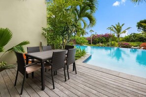 Eine Holzterrasse mit Tisch und Stühlen, einem Schwimmbecken und üppiger Bepflanzung, darunter Palmen und blühende Pflanzen.
