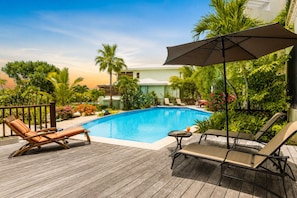 Un espace piscine avec des chaises longues et une table, entouré de verdure et d’un bâtiment.