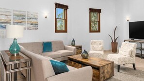 A living room with a sofa, armchairs, a coffee table, and a television. There are two windows with curtains, a lamp, and wall art.