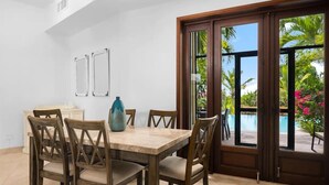 Un espace repas comprenant une table et des chaises, un vase, et une vue sur une piscine et la verdure, le tout visible à travers de grandes portes vitrées.