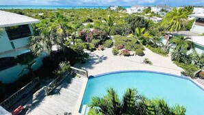 Ein Resort mit Swimmingpool, umgeben von üppiger Vegetation und Palmen, mit Blick auf den Ozean und weitere Gebäude des Resorts im Hintergrund.