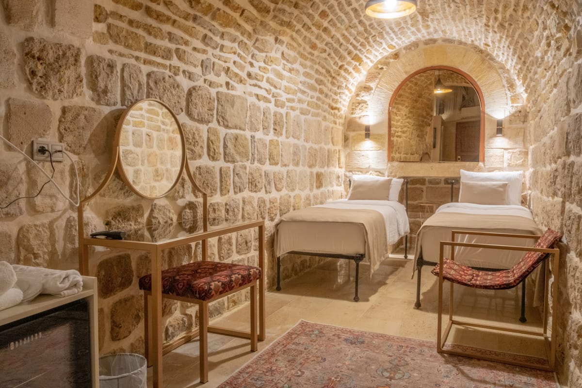 A stone-walled room with two beds, a wooden chair, a small table, and a mirror.