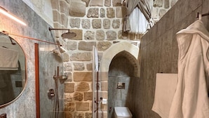 A stone-walled bathroom with a glass-enclosed shower, a toilet, and a mirror. - Ana Talia House (Mardin)