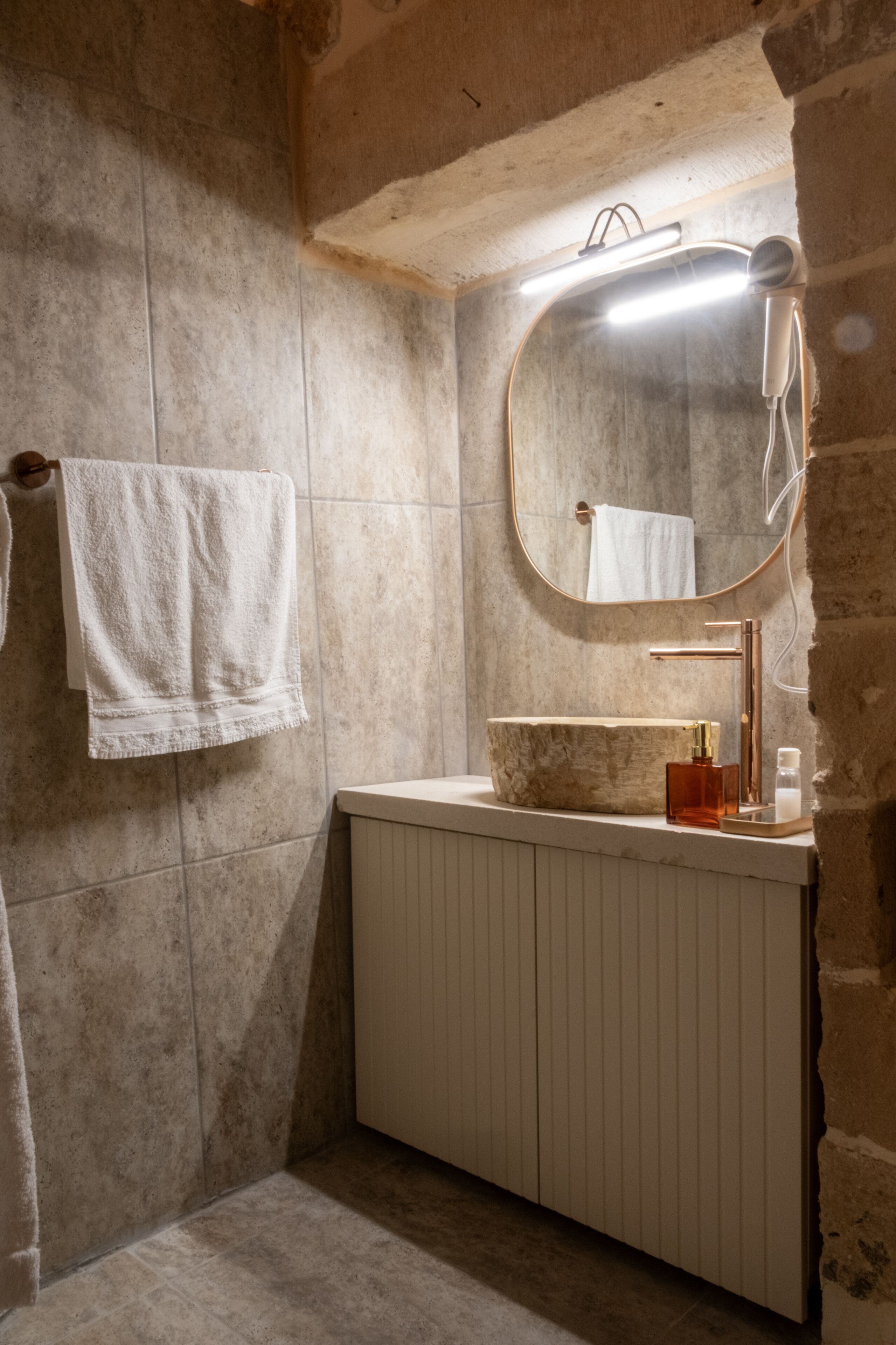 A bathroom with a stone wall, a white towel hanging, a mirror, a sink with a wooden countertop, and a towel rack.